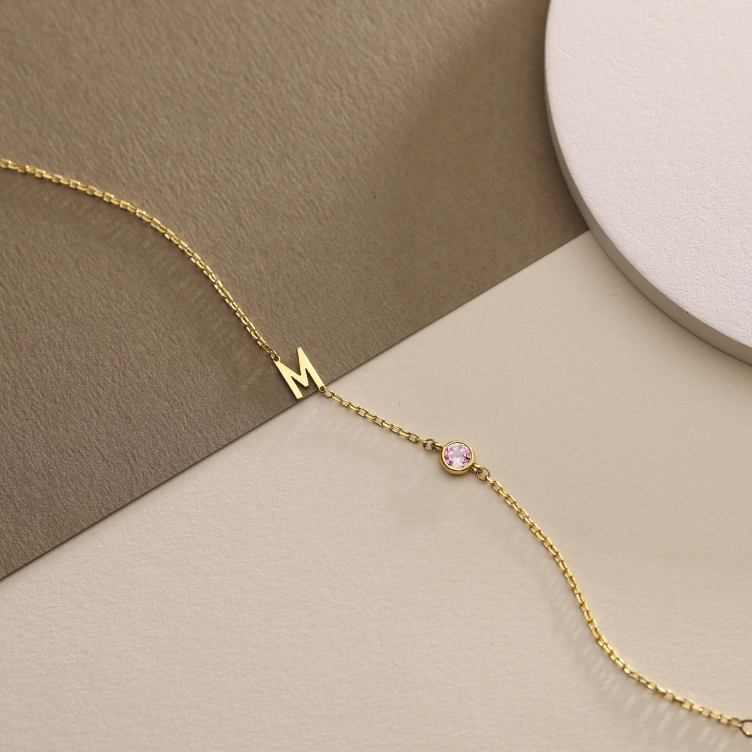 Gold M initial bracelet with one round pink birthstone detail on beige and taupe surfaces