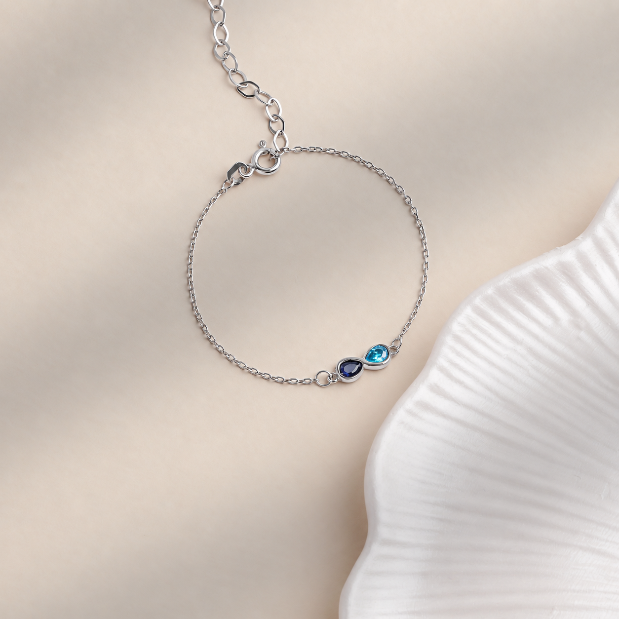Custom dual birthstone bracelet in solid white gold, with navy and aqua teardrop stones on a cream background beside a white ceramic dish