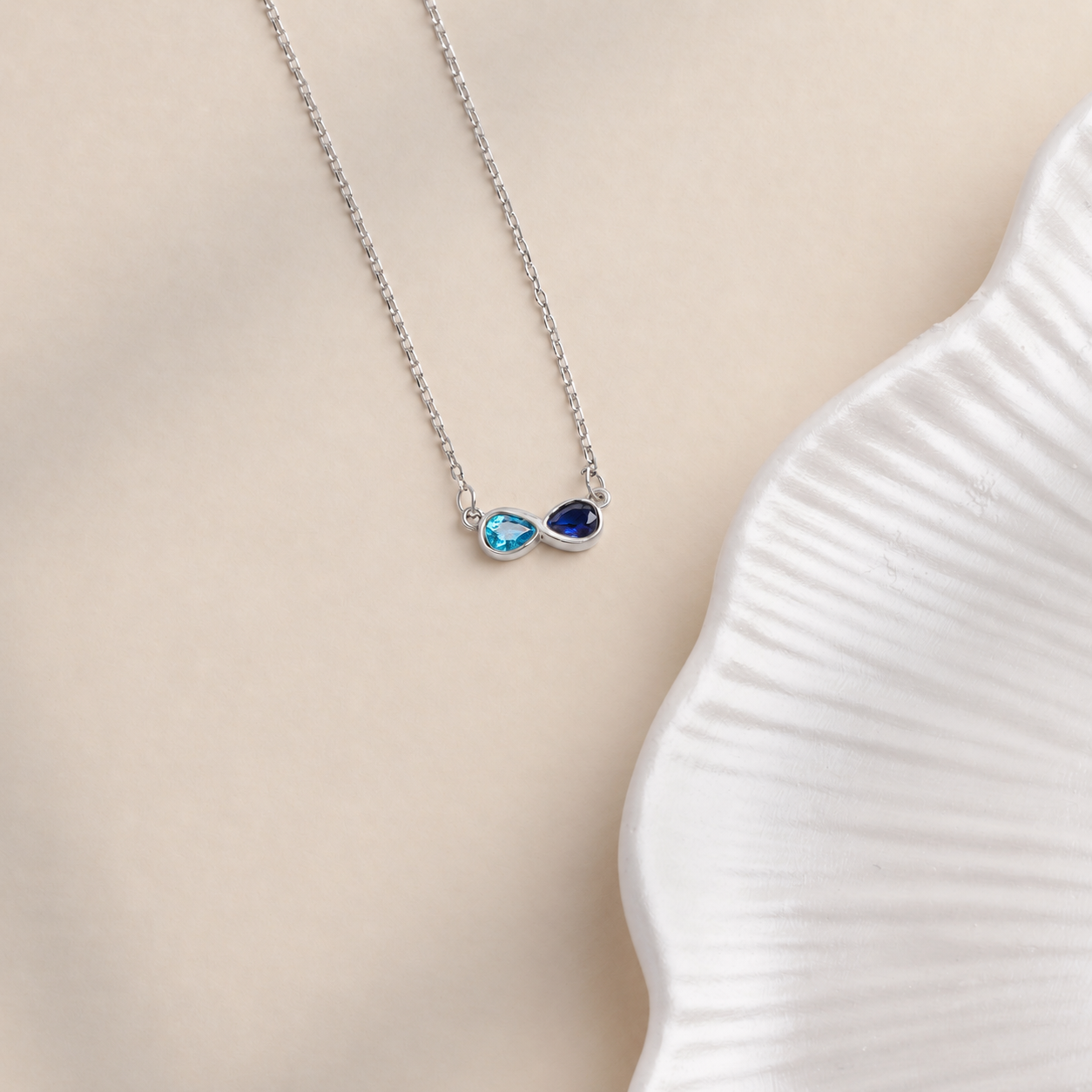 Custom dual birthstone necklace in solid white gold with light blue and navy pear-shaped gemstones on a cream background