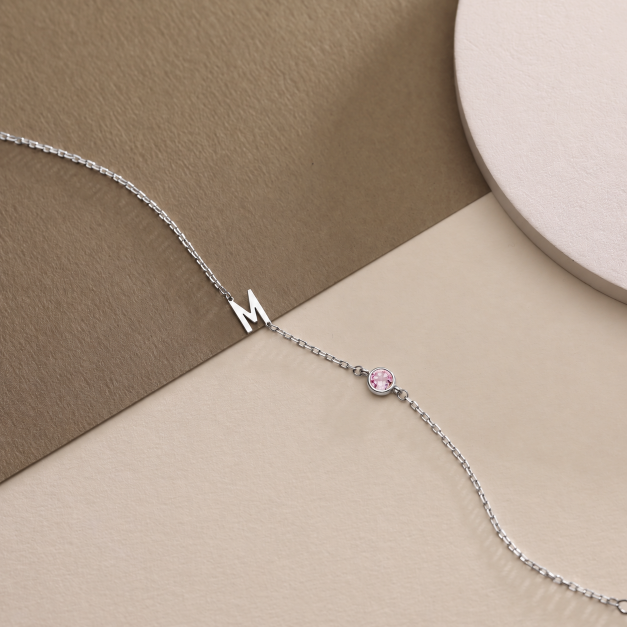 14K solid white gold M initial birthstone bracelet with a round pink gemstone on beige and taupe surfaces