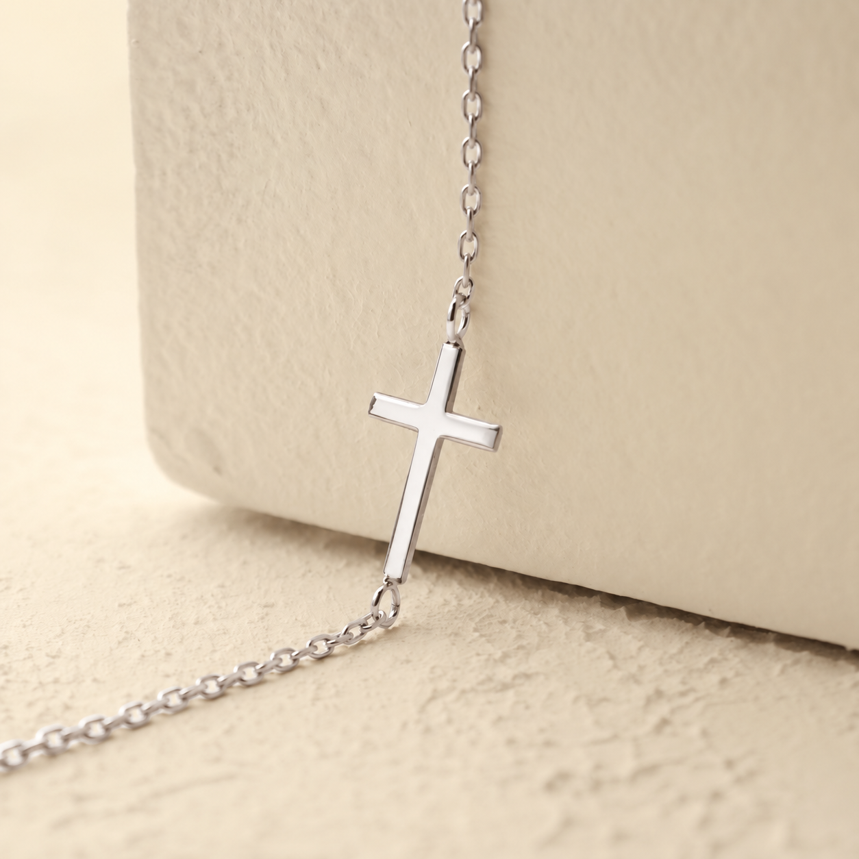 Dainty cross bracelet in solid white gold, close-up on a cream textured background