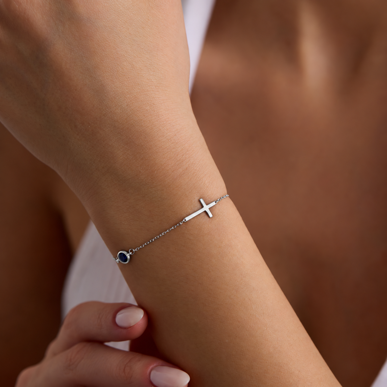 Sterling silver sideways cross bracelet with a round blue birthstone, worn on a wrist