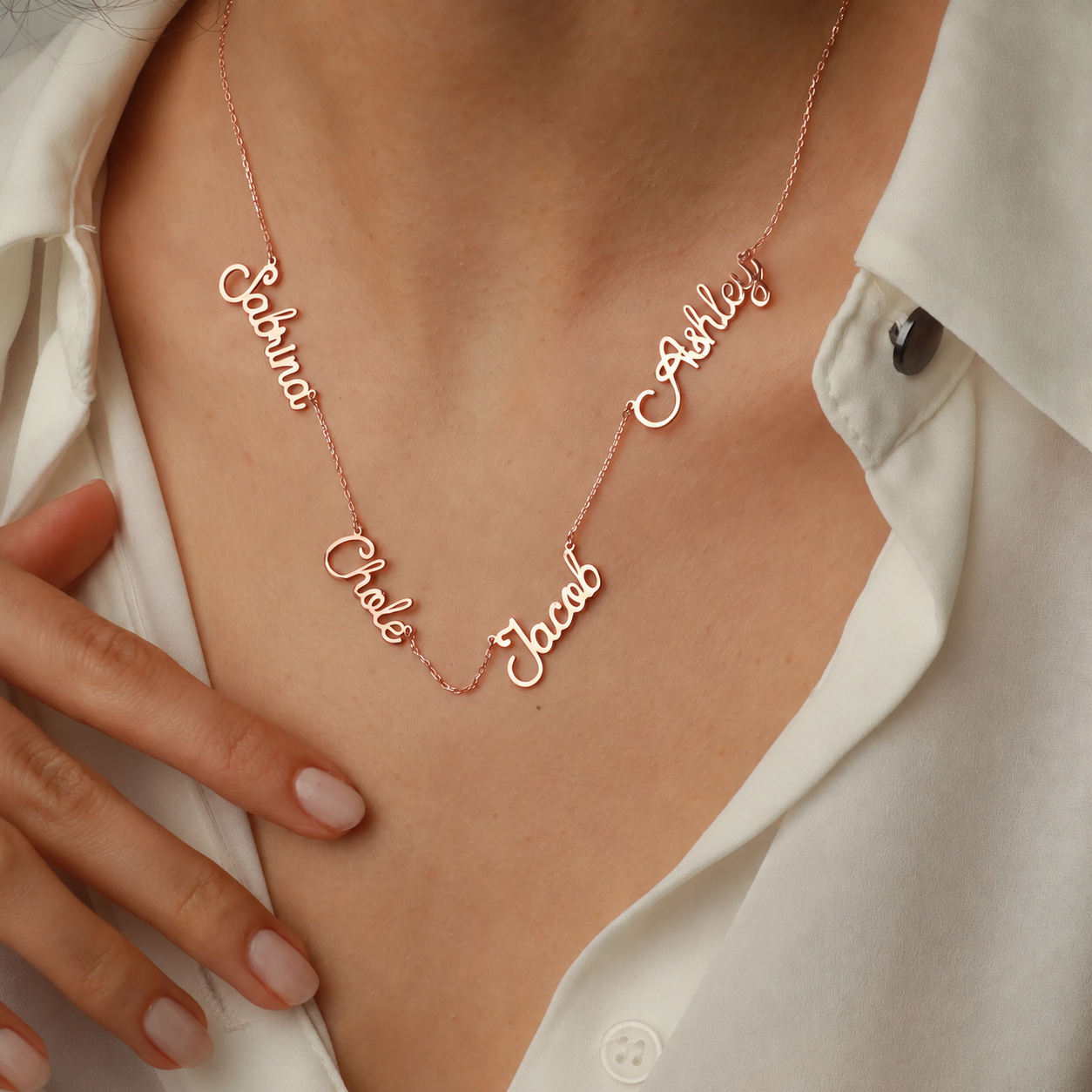 Family name necklace in rose gold, featuring four script name stations on a fine chain, worn with a white blouse.