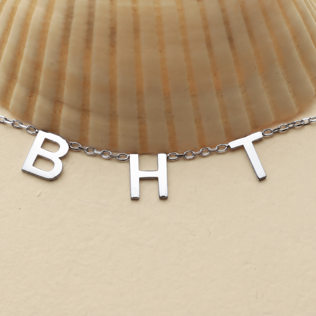 Initial letter necklace in solid white gold with B H and T charms beside a seashell on a cream background