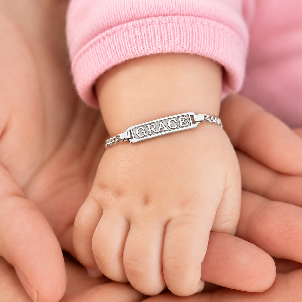Personalized baby name ID bracelet in 14K solid white gold, with a rectangular bordered GRACE nameplate and curb chain on a baby's wrist, supported by adult hands.