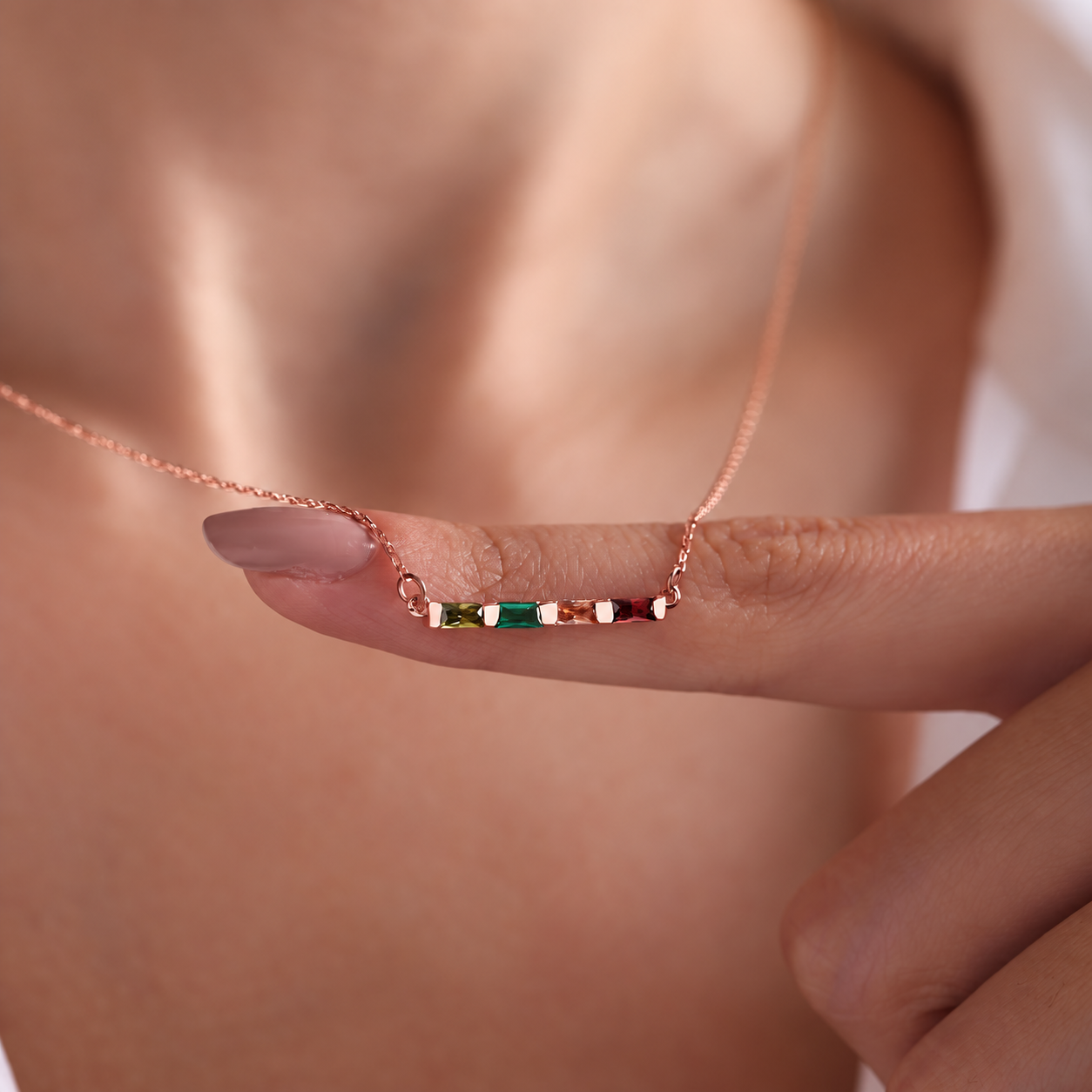 Personalized birthstone necklace in rose gold with four colourful baguette gemstones, held on a finger