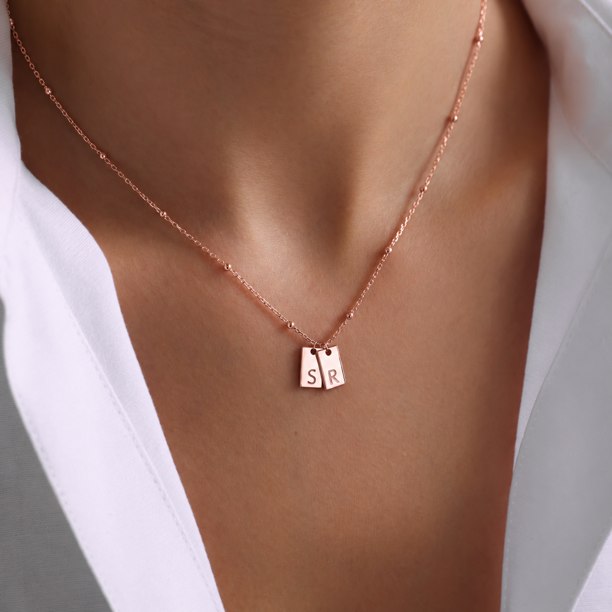 Personalized initial tag necklace in 14K rose gold filled with two rectangular pendants engraved S and R on a delicate beaded chain, worn with a white shirt.