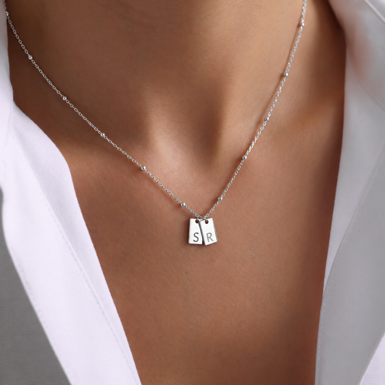 Personalized initial tag necklace in 925 sterling silver with two rectangular pendants engraved S and R on a delicate beaded chain, worn with a white shirt.