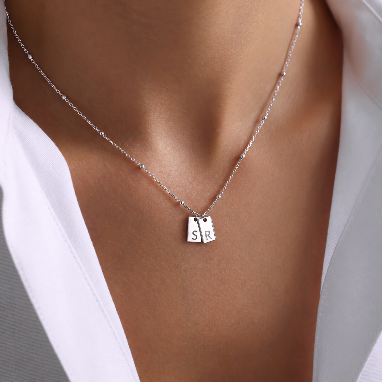 Personalized initial tag necklace in 14K solid white gold with two rectangular pendants engraved S and R on a delicate beaded chain, worn with a white shirt.