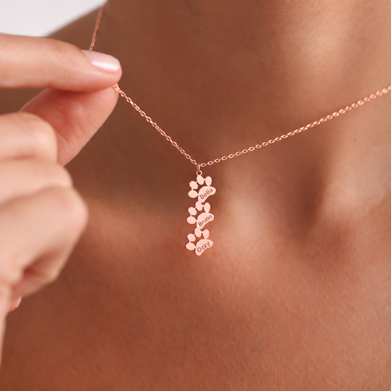 Personalized pet loss necklace in 14K rose gold filled, with three connected vertical paw shapes engraved Bella, Bruno and Ozzy, held against the neckline.