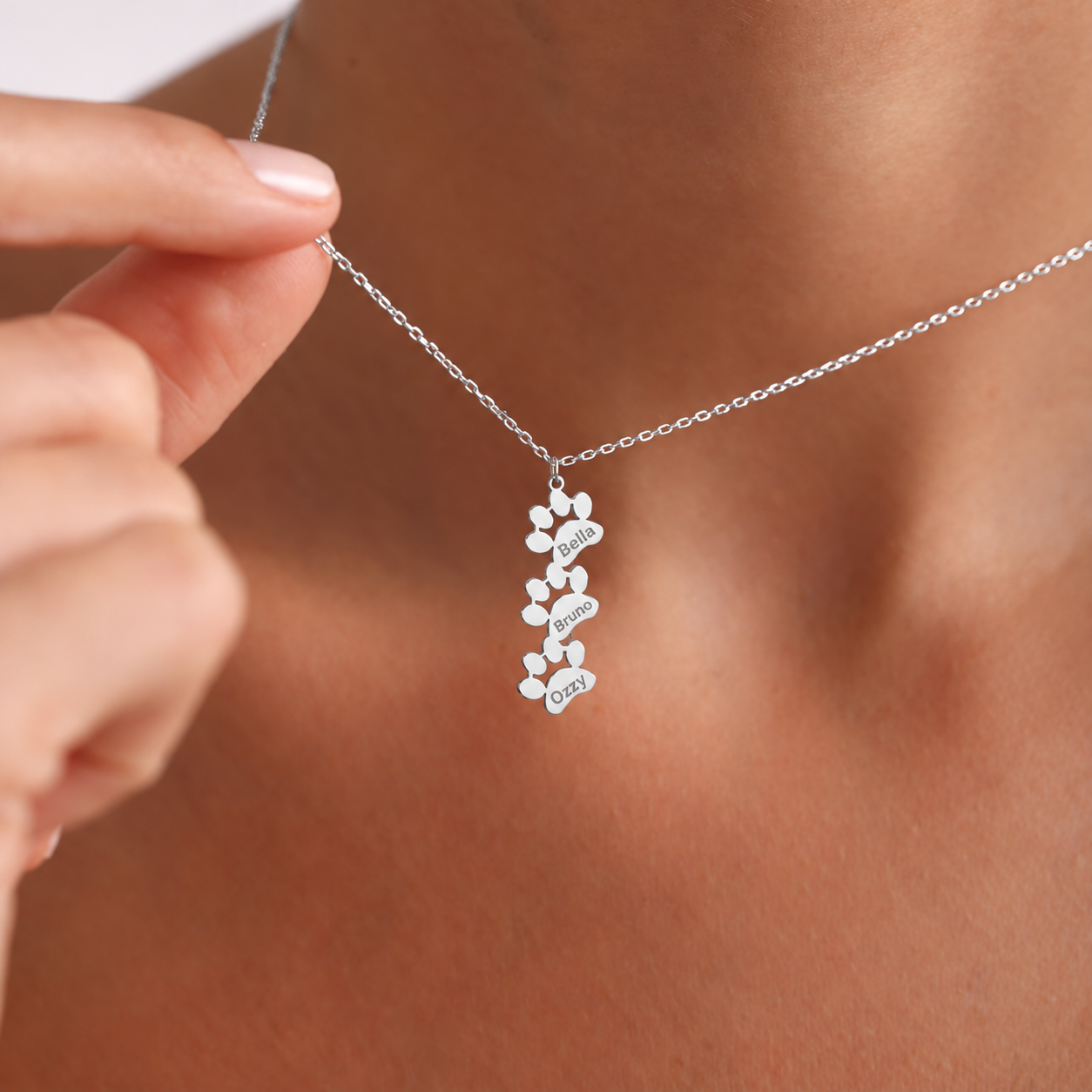 Personalized pet loss necklace in 925 sterling silver, with three connected vertical paw shapes engraved Bella, Bruno and Ozzy, held against the neckline.