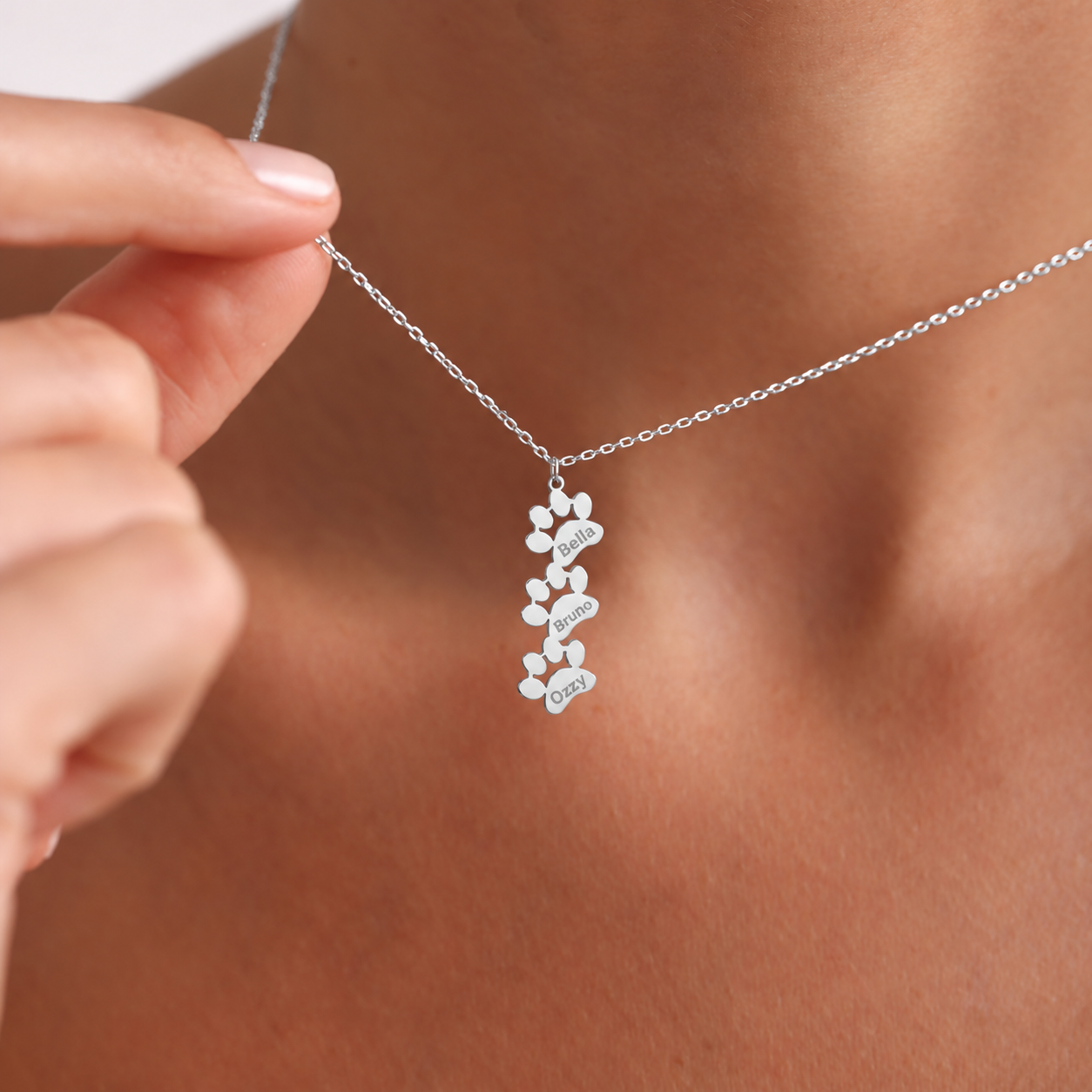 Personalized pet loss necklace in 14K solid white gold, with three connected vertical paw shapes engraved Bella, Bruno and Ozzy, held against the neckline.
