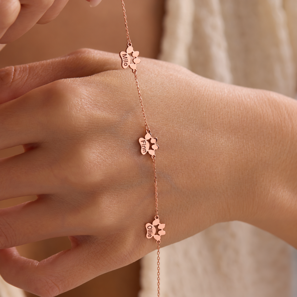 Personalized pet paw bracelet in 14K rose gold filled with three paw-shaped stations engraved Milo, Finn and Ollie, displayed across a hand.