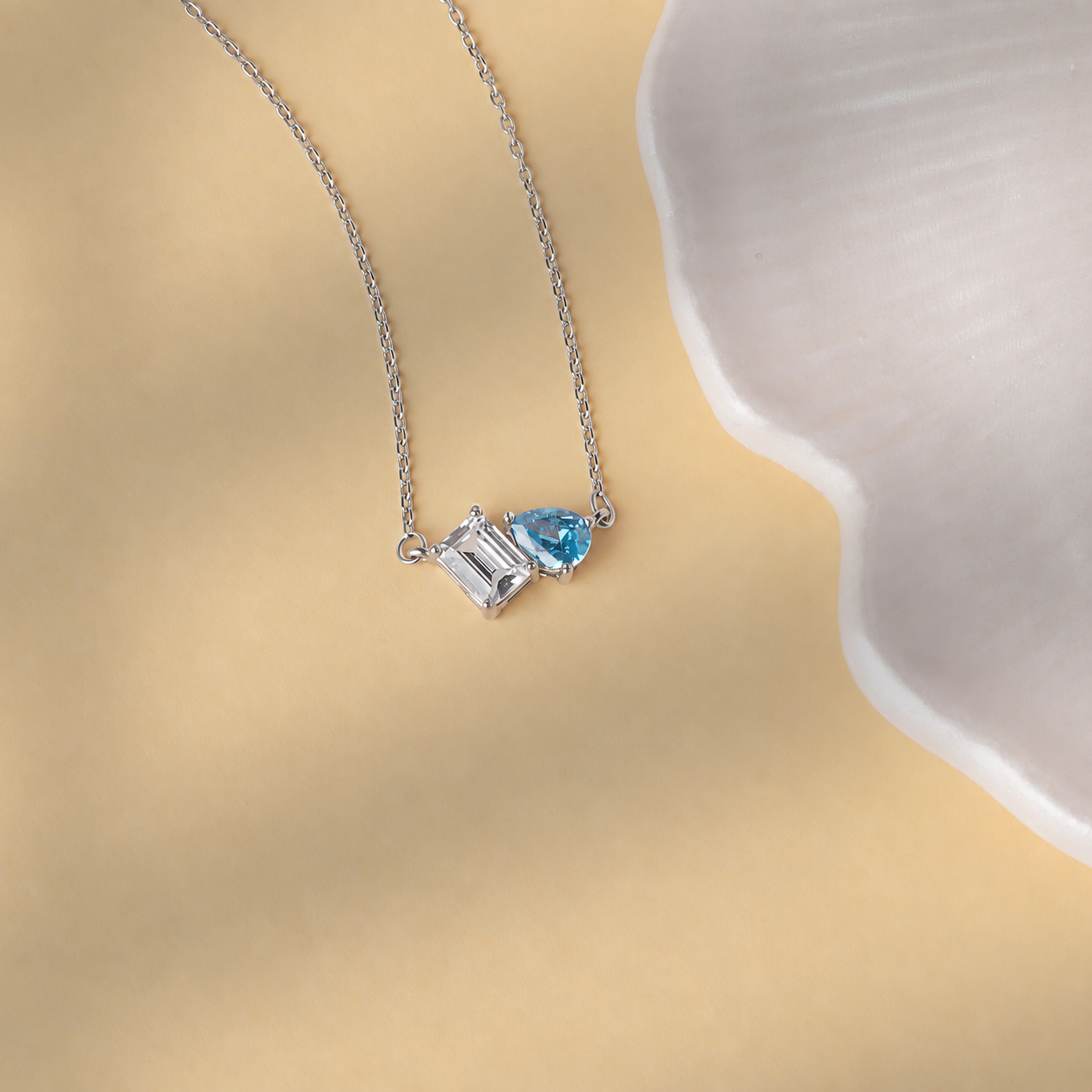 Toi et Moi birthstone necklace in solid white gold with clear rectangular and blue pear-shaped gemstones on a cream background
