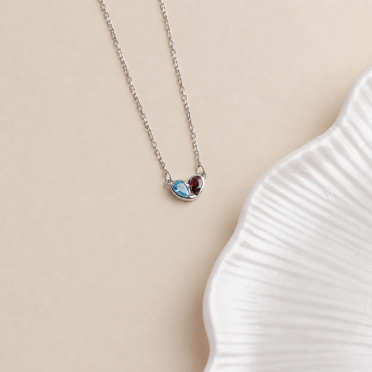 Two birthstone heart pendant necklace in solid white gold with blue and red gemstones on a cream background
