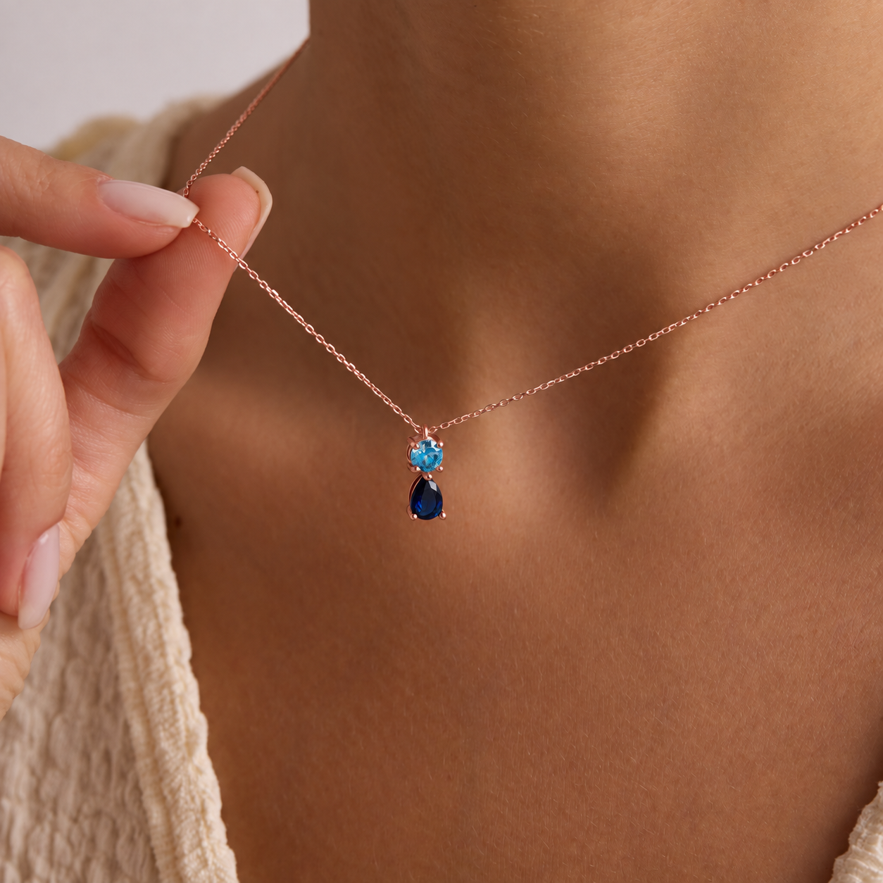 Two birthstone pendant necklace in rose gold with a round light blue stone above a navy pear-shaped stone, worn on the neck