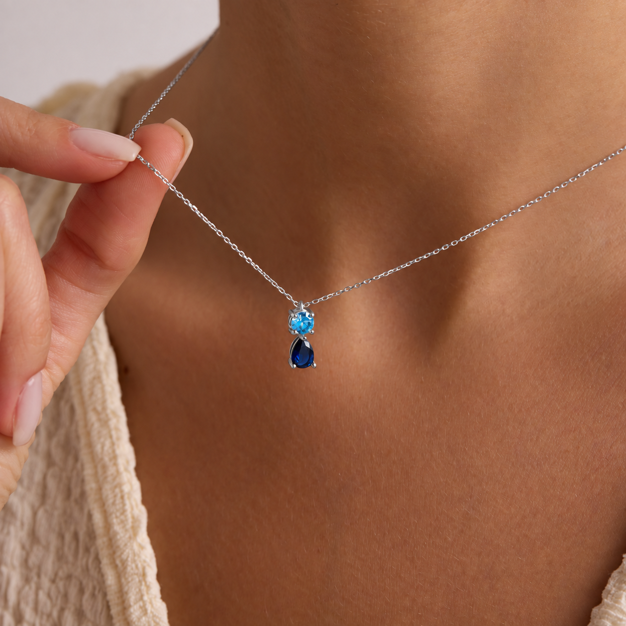 Two birthstone pendant necklace in sterling silver with a round light blue stone above a navy pear-shaped stone, worn on the neck