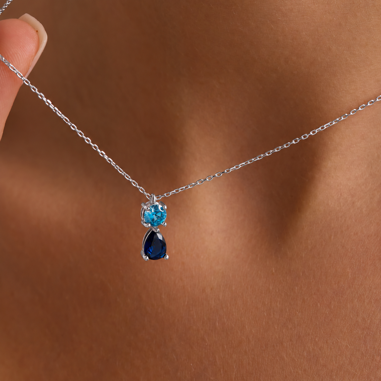 Two birthstone pendant necklace in solid white gold with a round light blue stone above a navy pear-shaped stone, close-up